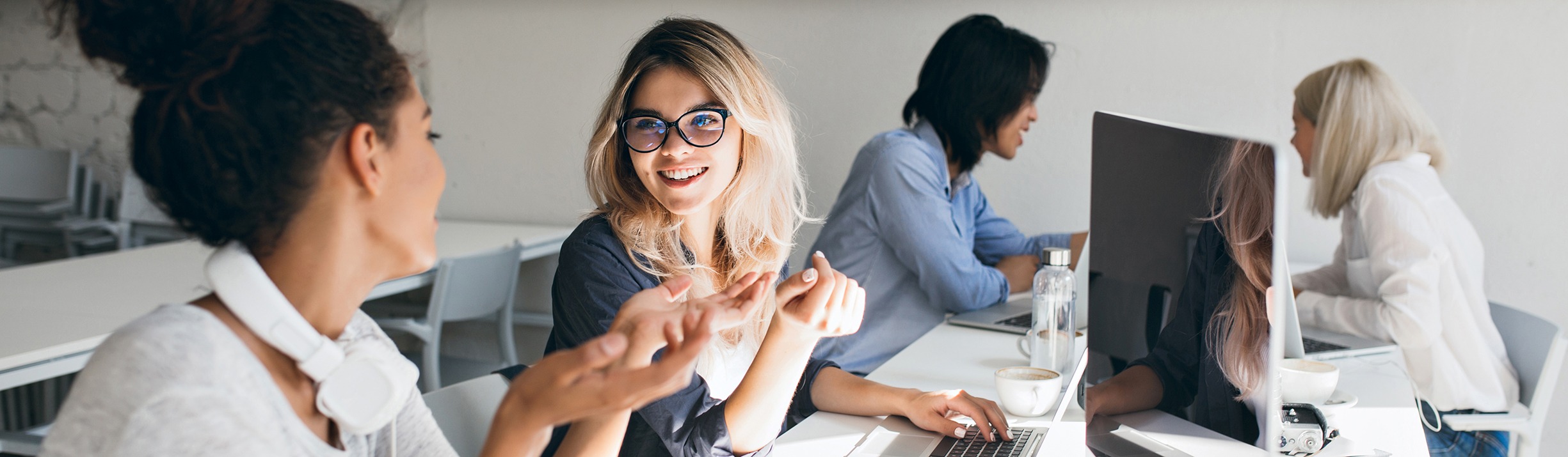 Curly mulatto girl in gray t shirt explains something to blonde female friend. indoor portrait of international students with laptops preparing for test together..